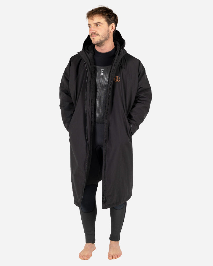 A person wearing a green and black Fourth Element Tidal Robe is embracing another person who is wearing an orange garment, standing on a beach with the ocean in the background.